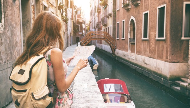 Jeune femme randonneuse avec un sac à dos beige, consultant une carte au bord d'un canal calme de Venise au soleil couchant.