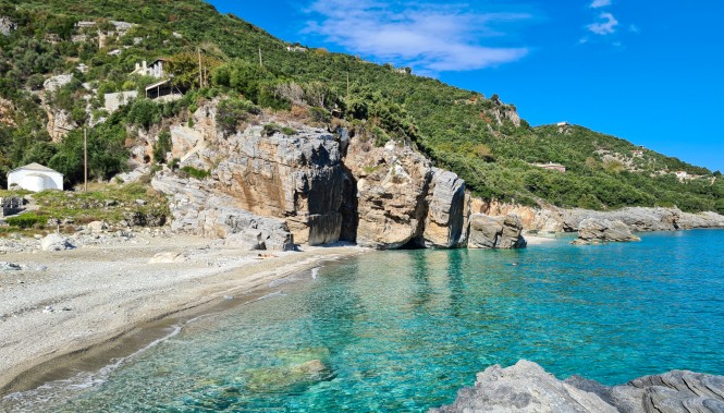 Crique de galets en Grèce avec eaux turquoise, falaises rocheuses et formation de grotte en bord de mer.