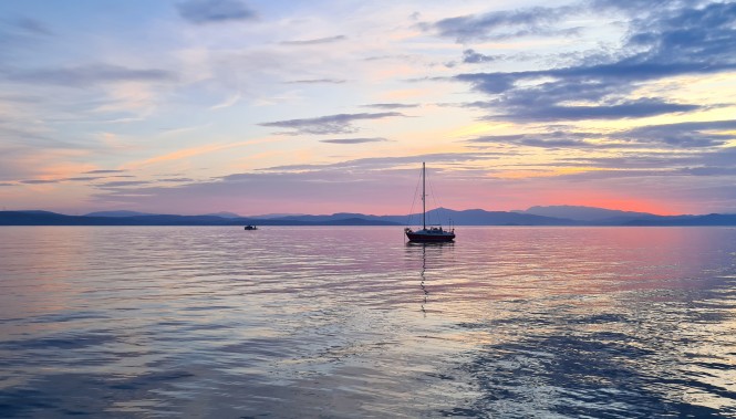 Coucher de soleil aux couleurs chaudes (rose, orange, violet) sur un voilier naviguant en mer Égée, Grèce.